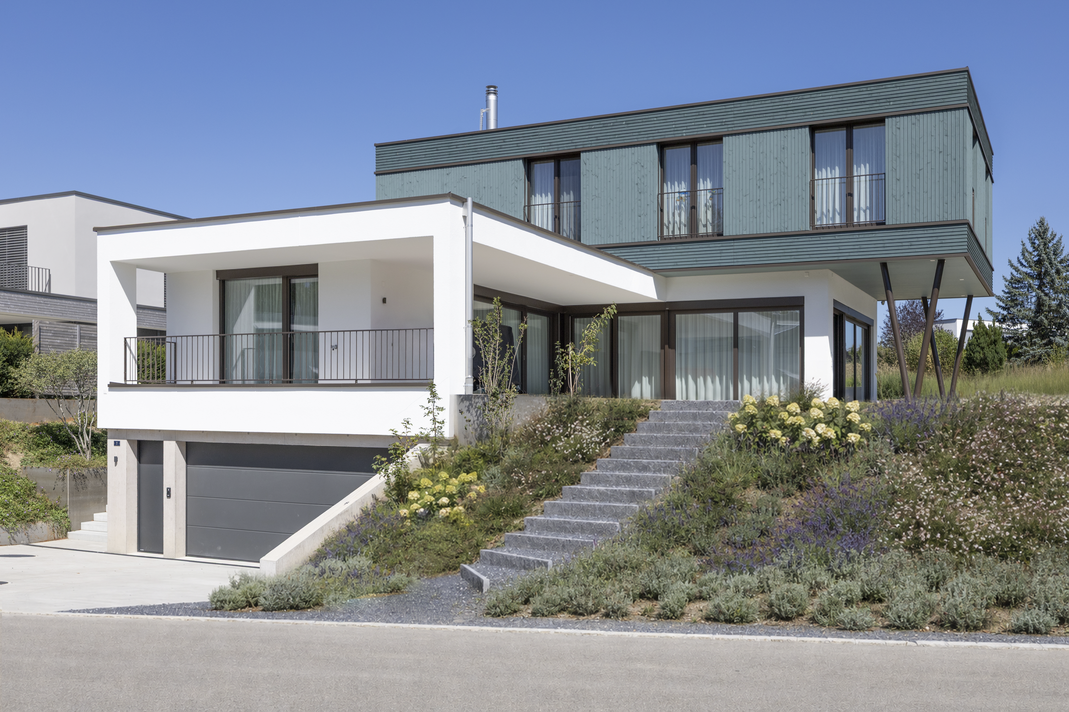 Südwest-Ansicht Ein Haus ist weit mehr als ein Gebäude – es ist ein Ort zum Ankommen, Leben und Wohlfühlen. Farben und Materialien spielen dabei eine zentrale Rolle. Natürliche Holzoberflächen, Fassadenelemente und fein abgestimmte Farbakzente erzeugen eine warme, einladende Atmosphäre. Ein Zuhause, das Ästhetik, Funktion und Nachhaltigkeit vereint – ein Ort, an dem Farbe, Form und Raum zu einer harmonischen Einheit verschmelzen.
